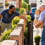 A man pauses his gardening to cheerfully hold a gate open for an older neighbour carrying boxes, demonstrating kindness and putting faith into practice.