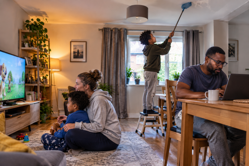 An older boy stands on a stool dusting the ceiling while his younger sibling enjoys television with their mother and their father works on a laptop nearby, illustrating the unequal expectations sometimes placed on firstborn children.