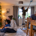 An older boy stands on a stool dusting the ceiling while his younger sibling enjoys television with their mother and their father works on a laptop nearby, illustrating the unequal expectations sometimes placed on firstborn children.
